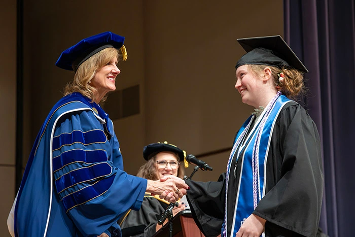 A graduate shakes hands with President Mazachek and waves to the crowd as they walk across the stage.