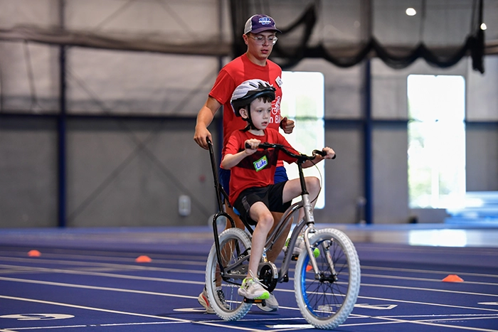 Washburn University kinesiology student Alex Broxterman walks behind an iCan Bike participant as the child rides a two-wheel bicycle with a training support bar during an indoor iCan Bike camp.