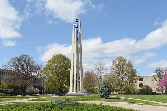 Washburn University's bell tower rises above the campus lawn, surrounded by trees and walkways under a blue sky.