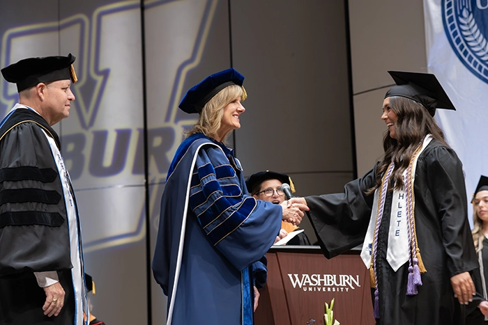 Washburn University President Dr. JuliAnn Mazachek shakes hands with a smiling graduate wearing honor cords and a student-athlete stole during commencement ceremonies on stage. Faculty members and the Washburn University logo are visible in the background.