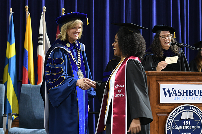 Washburn University President JuliAnn Mazachek shakes hands with a graduate wearing a cap, gown and Class of 2026 stole during commencement.