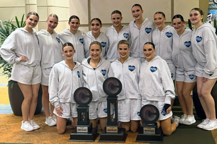 The Dancing Blues pose together indoors, wearing matching light-colored hoodies, shorts, and white sneakers. They smile at the camera while arranged in two rows, with some kneeling in front. Three large trophies are placed on the floor in front.