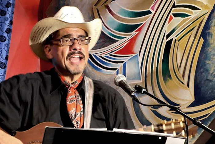 Man singing and playing guitar in front of an intricate background.  